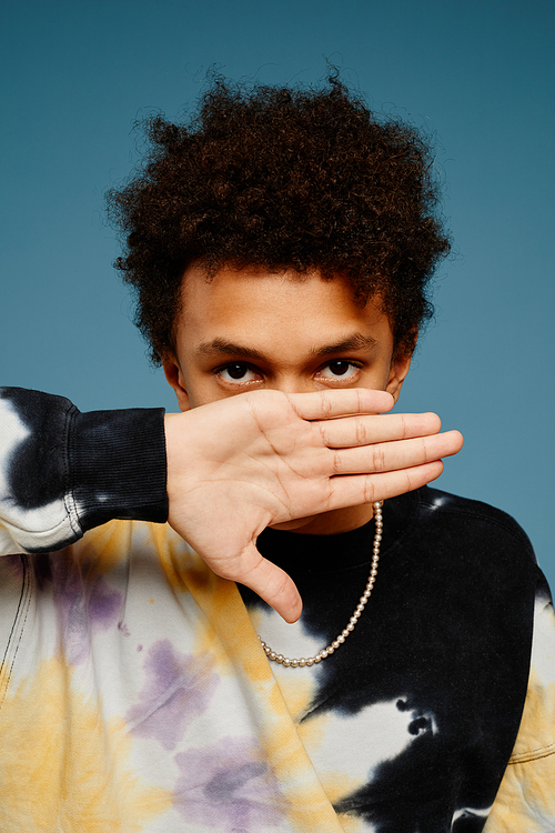 Vertical portrait of black teenage boy wearing tie dye shirt and hiding face over blue background