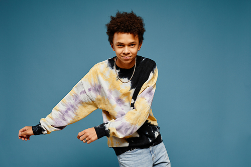Minimal portrait of black teenage boy wearing tie dye shirt and dancing over blue background, copy space