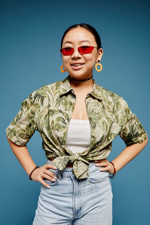 Vertical portrait of Asian teenage girl wearing trendy colored sunglasses over blue background