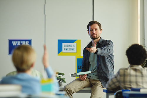 Portrait of bearded male teacher asking questions during class with children raising hands, copy space