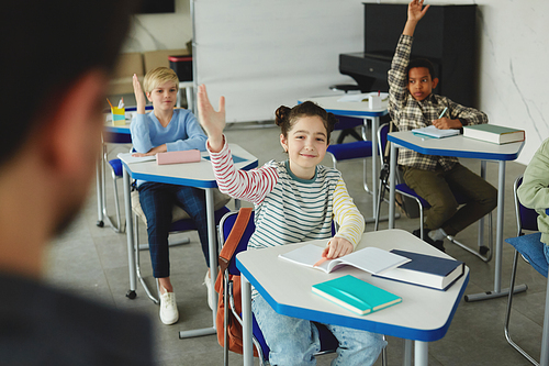 Portrait of smiling young girl raising hand in class and looking at teacher, copy space