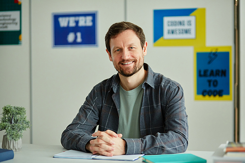 Front view portrait of smiling bearded teacher looking at camera while sitting at desk in school classroom against colorful posters