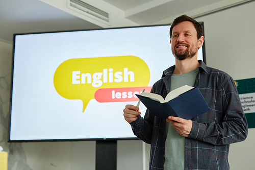 Waist up portrait of smiling male teacher holding book while standing against screen with English lesson text, copy space