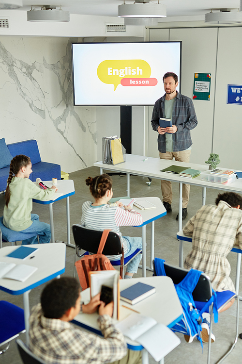 Vertical portrait of diverse group of children studying in school classroom with male teacher watching during English lesson