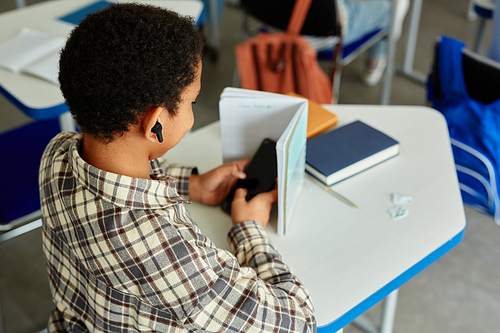 High angle view at young black child listening to music with wireless earphones during school class and hiding phone, copy space