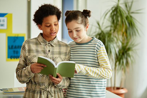 Waist up portrait of two girls answering questions in school in front of classroom and holding book