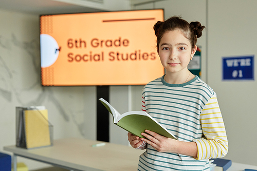 Waist up portrait of cute schoolgirl in striped shirt looking at camera and holding book while standing in classroom, copy space