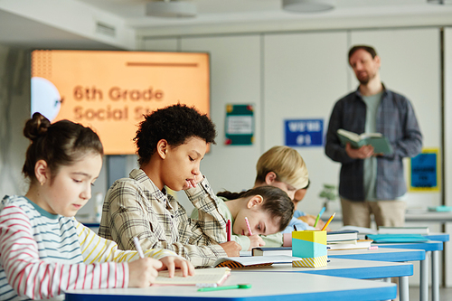 Side view at diverse group of children sitting in row and taking test in school classroom with teacher in background