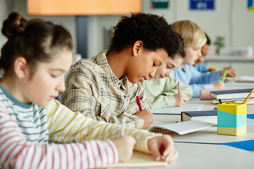 Side view portrait of young black girl taking test in school classroom with group of children sitting in row