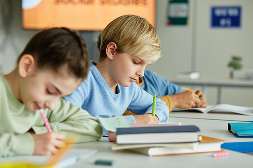 Side view portrait of young blonde boy taking test in school classroom with group of children sitting in row