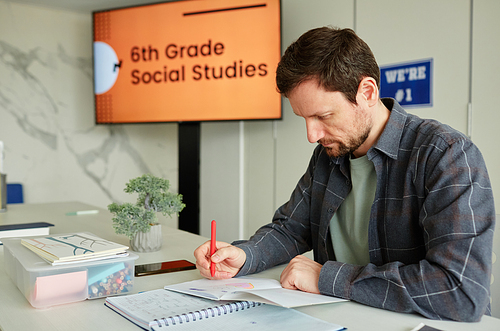 Portrait of male teacher sitting at desk in school classroom and grading childrens homework