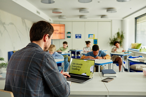 Back view at male teacher using laptop at desk in classroom with group of children studying in background, copy space