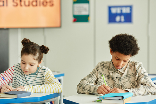 Front view at two children sitting at desk in classroom and writing in notebooks while studying in school