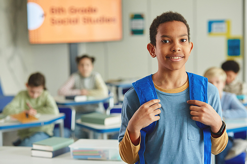 Waist up portrait of black young schoolboy with backpack looking at camera while standing in classroom, copy space