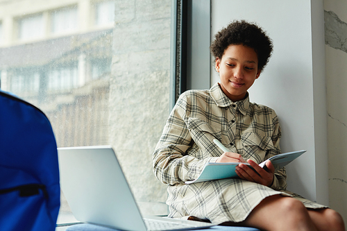 Portrait of smiling African American kid writing in notebook while sitting on window in school with laptop, copy space