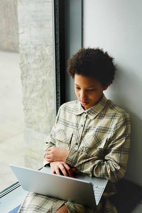 Vertical portrait of young black girl using laptop and watching videos while sitting by window in school