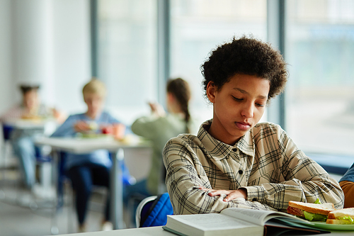 Portrait of young black girl reading book at lunch break in school canteen, copy space