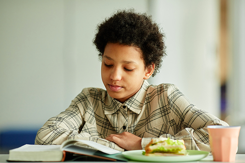 Front view portrait of young African American girl reading book during lunch break in school canteen