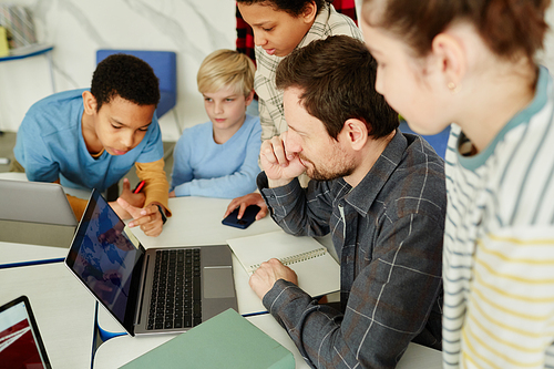 High angle portrait of male teacher with diverse group of kids using laptop in school