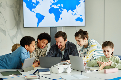 Front view portrait of smiling male teacher with diverse group of children using laptop together in modern school classroom