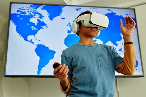 Waist up portrait of young black boy using VR technology in school classroom with geography map in background, copy space