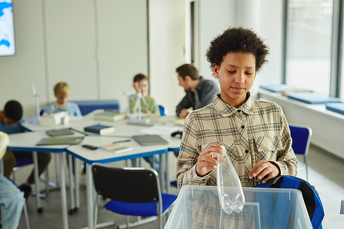 Waist up portrait of young black girl using waste sorting bins in school and putting plastic bottle in, copy space