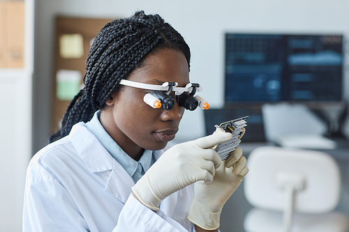 Portrait of young African American woman wearing magnifying visor and inspecting hardware part in engineering laboratory