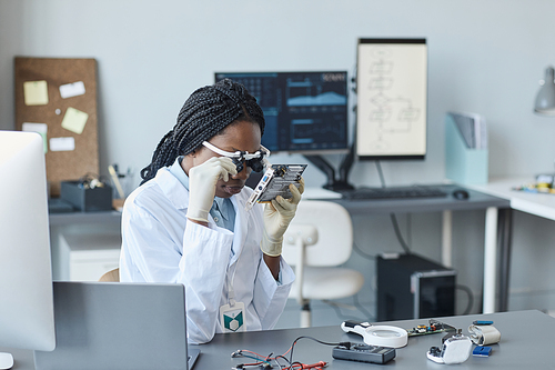 Portrait of young black woman wearing magnifying visor and inspecting hardware part in engineering laboratory