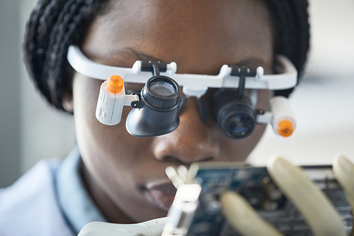 Close up of young black woman wearing magnifying visor and inspecting hardware part in engineering laboratory