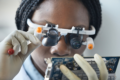 Close up of black female scientist wearing magnifying glasses and inspecting hardware part in engineering lab