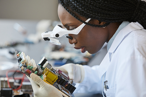 Side view portrait of black female scientist wearing magnifying glasses and inspecting electronic parts in quality control lab