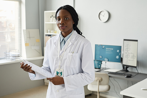 Waist up portrait of young black woman wearing lab coat while working in research facility and looking at camera, copy space