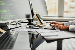 Close-up of female programmer typing code on keyboard from smartphone while sitting at table in front of computer monitors at office