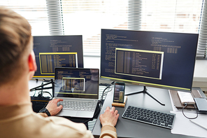 Rear view of young IT specialist connecting laptop with computer while typing data on keyboard sitting at his workplace