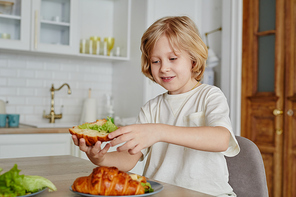 Portrait of smiling blonde boy enjoying breakfast in morning and eating sandwich in kitchen interior, copy space