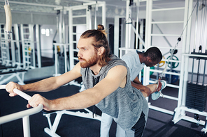 Young patient concentrating on his stretching exercises on sport equipment in gym together with doctor helping him