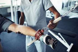 Close-up of patient training his leg on sport equipment together with nurse supporting him during their exercises in gym