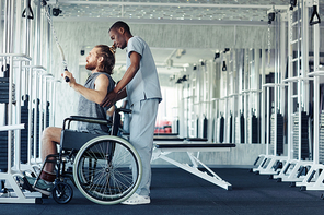 Young patient with disability sitting in wheelchair and training his back on equipment with the help of doctor