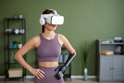 Young woman with prosthetic arm doing exercises in VR glasses in the room