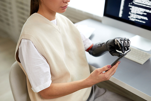 Close-up of young girl with prosthetic arm typing codes on smartphone while working at her workplace at home