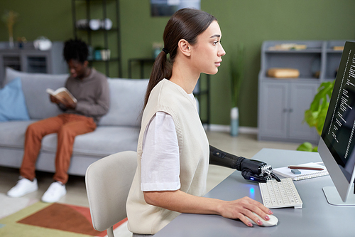 Serious young girl with disability working with codes on computer at her workplace at home with man reading book in background