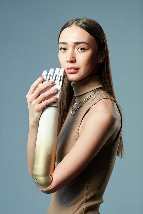 Portrait of young beautiful woman with prosthetic arm looking at camera standing against blue background