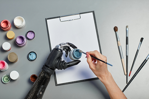 High angle view of woman with prosthetic arm painting with paints and brush on paper at table