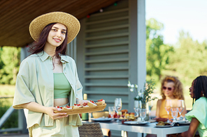 Waist up portrait of young woman holding tray of snacks looking at camera while enjoying outdoor party with friends in Summer, copy space