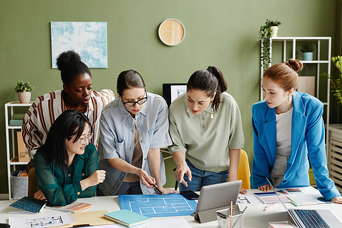 Group of female designer standing at table with blueprint and discussing it in team at office