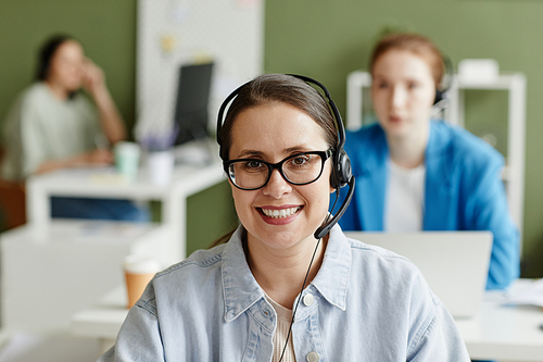 Portrait of young call centre agent in headphones and eyeglasses smiling at camera while working at office