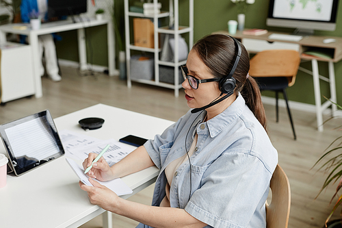 Young businesswoman in headphones making notes while talking to customer during online call on tablet pc sitting at her workplace