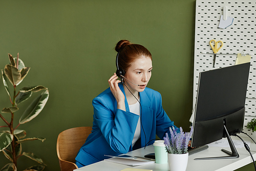 Young businesswoman in headset sitting at table in front of computer monitor and talking to her colleagues online