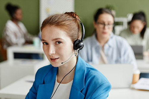 Portrait of young call center in headphones looking at camera while working at call center with her colleagues in background