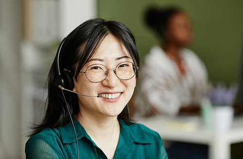 Portrait of Asian young operator in headphones and eyeglasses smiling at camera during her work at call centre